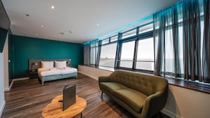 Ein modernes Hotelzimmer mit einem großen Bett, einer Couch, einem Schreibtisch mit einem Laptop und Meerblick durch große Fenster.