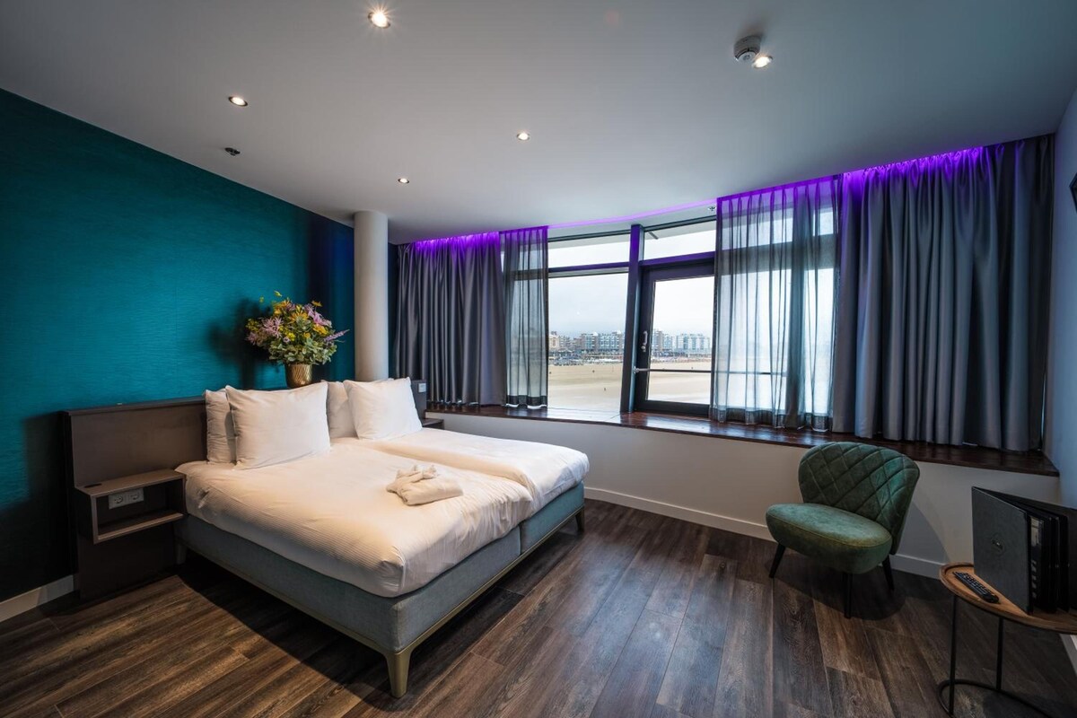 Una habitación de hotel moderna con una cama grande, vista a la ciudad e iluminación morada.