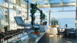 Ein Frühstücksbuffet mit verschiedenen Speisen, einem Laptop und Meerblick durch große Fenster.