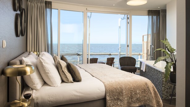 Ein Schlafzimmer mit einem großen Bett, einem Nachttisch mit Lampe, einem Weinglas und Blick auf das Meer durch große Fenster.