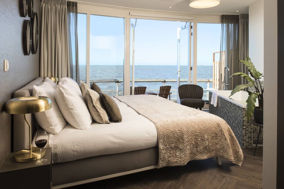 A bedroom with a large bed, a nightstand with a lamp, a glass of wine, and a view of the sea through large windows.