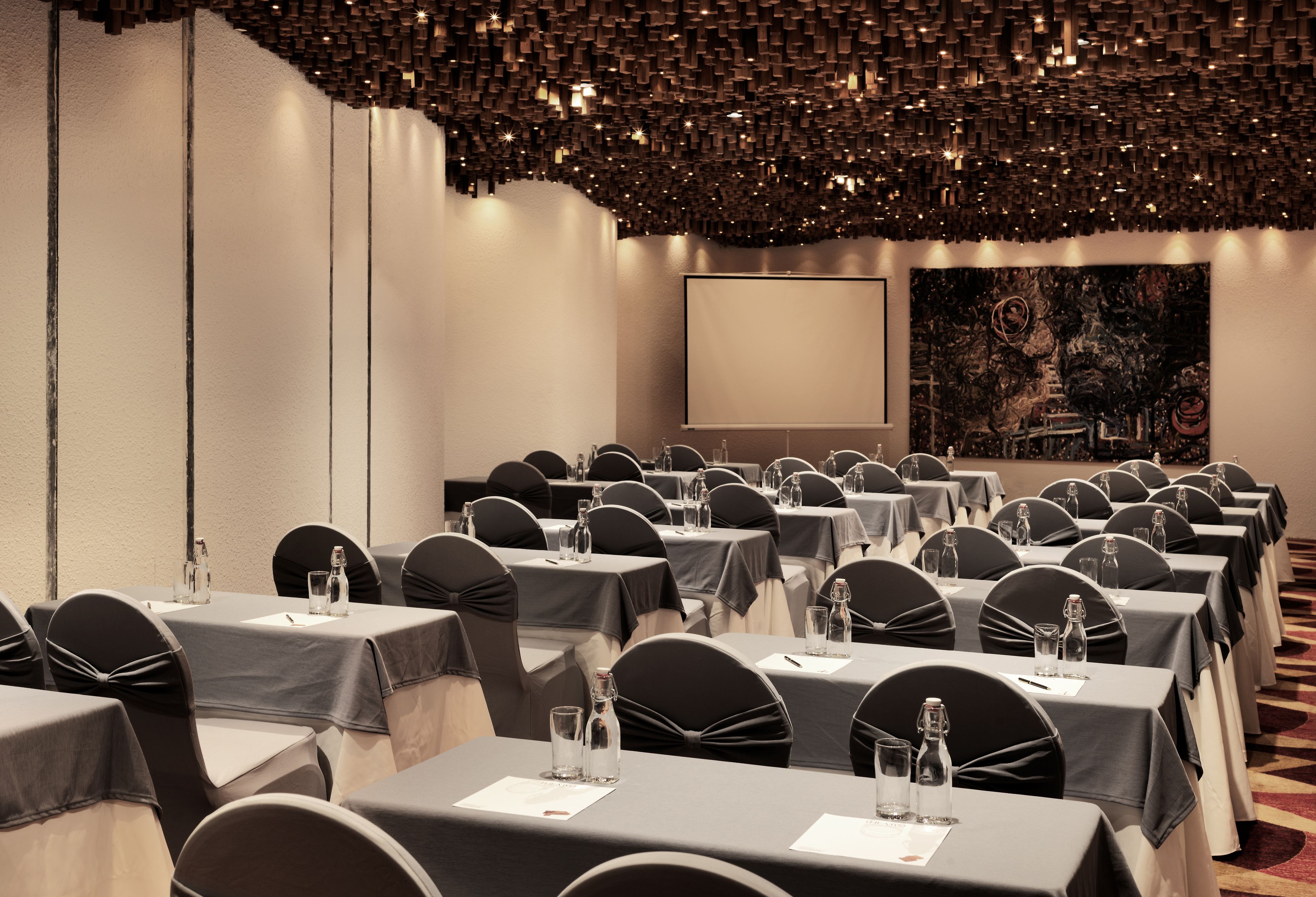 Ein Konferenzraum mit Reihen von Tischen, schwarzen Stühlen und einer Projektionsleinwand.