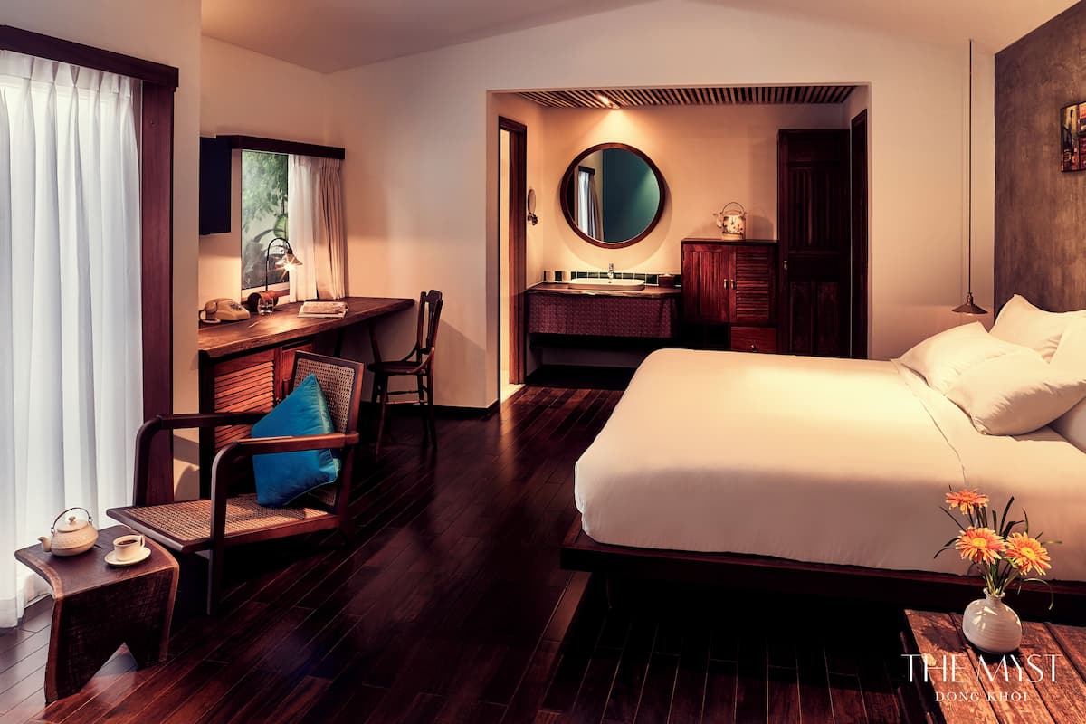 Une chambre d’hôtel avec un grand lit, un bureau avec une chaise, un miroir rond, une coiffeuse avec un lavabo et une petite table sur laquelle se trouvent une théière et une tasse.
