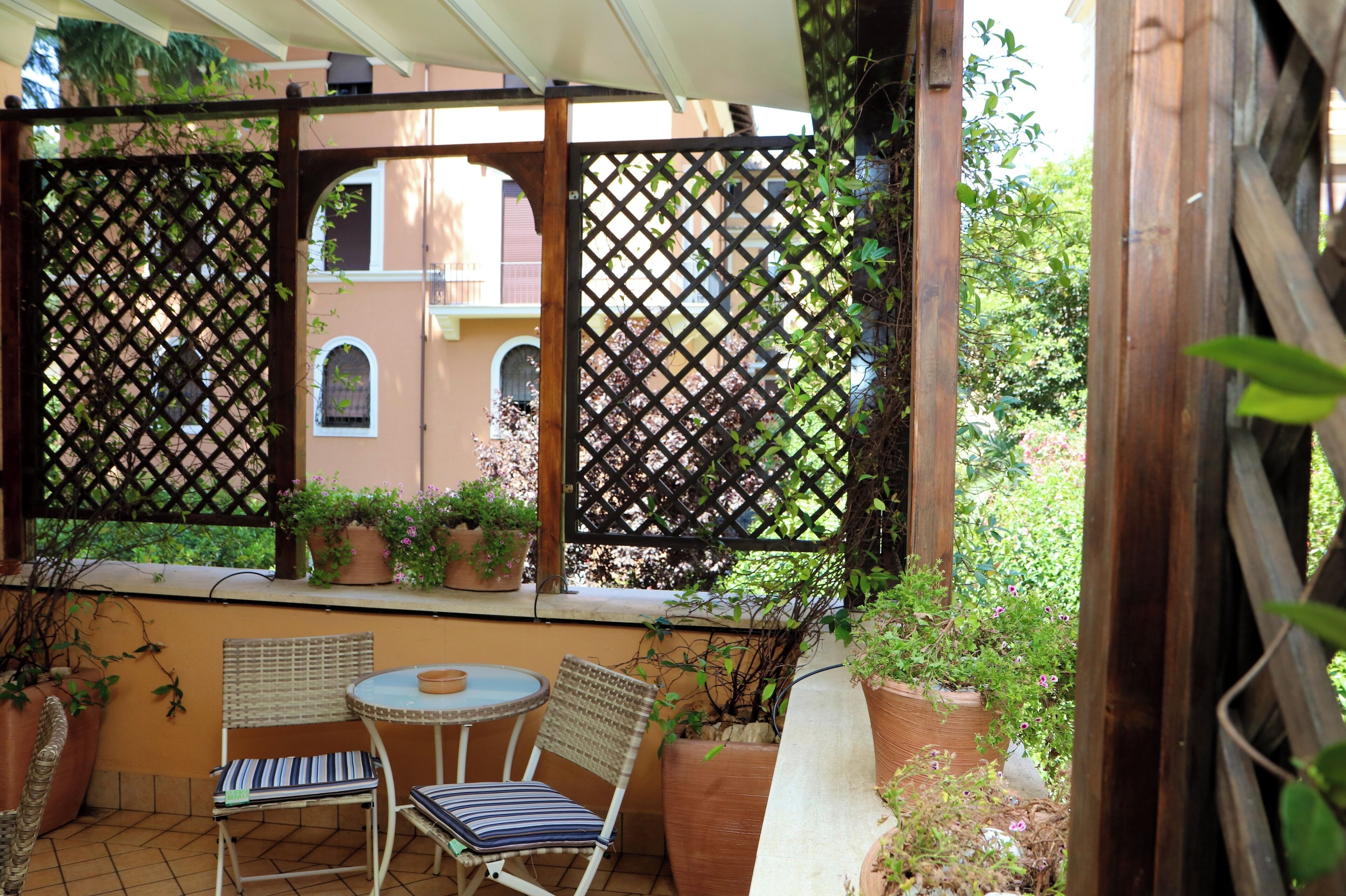 Une terrasse couverte avec deux fauteuils en osier et une petite table ronde, entourée de plantes en pot et d’une clôture en treillis.