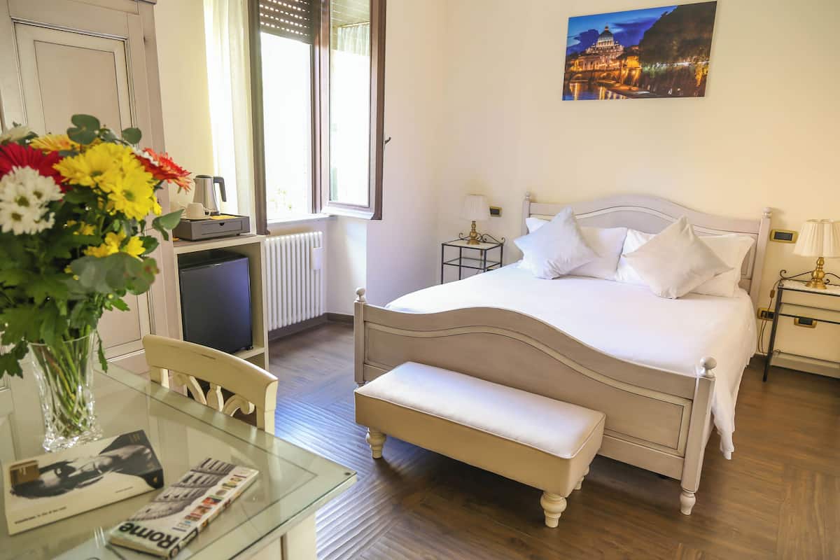 A hotel room with a bed, bedside tables, a chair, a glass table with a vase of flowers, a radiator, and a window with curtains.