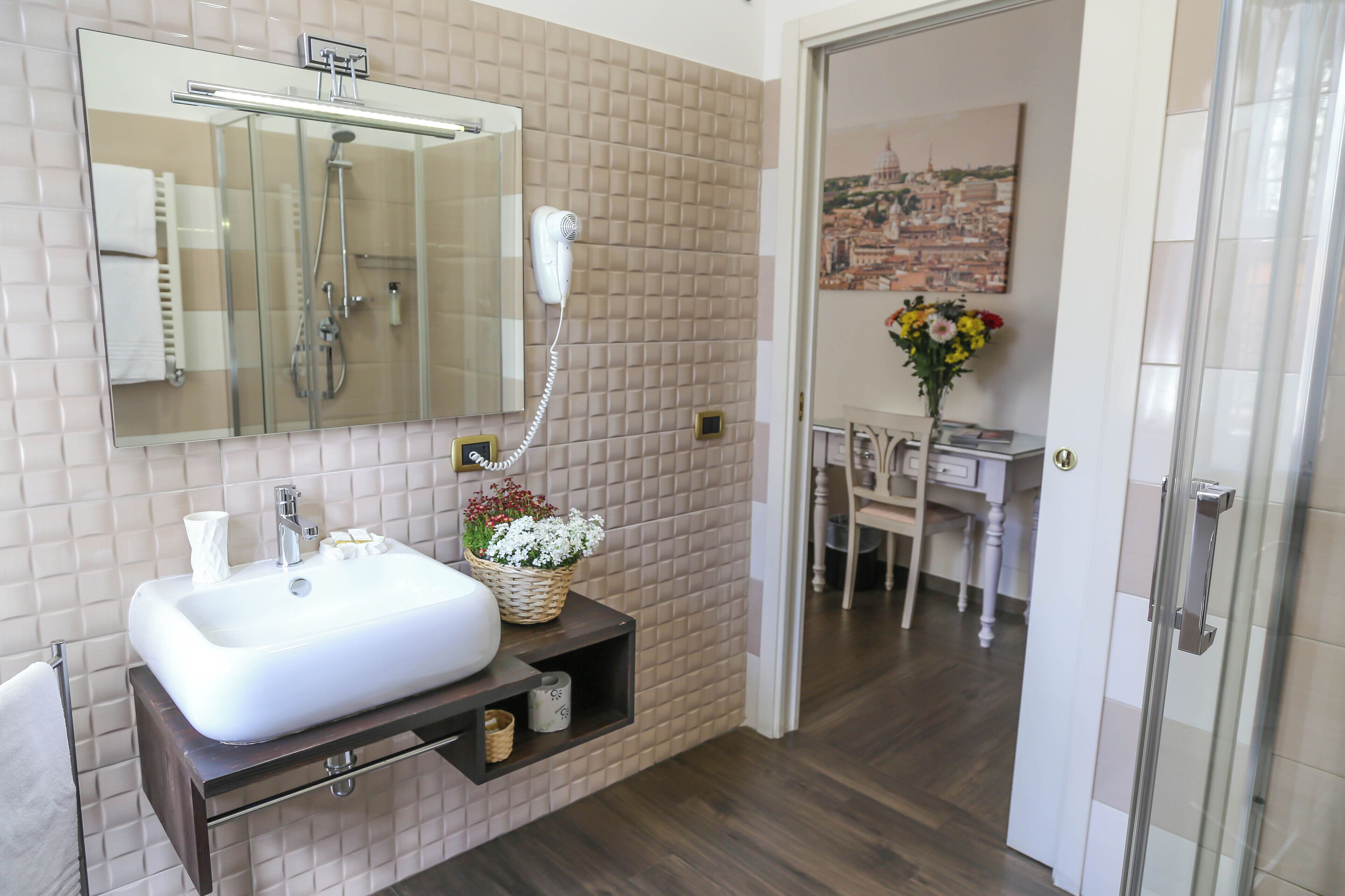 A bathroom with a white sink, a mirror, and a shower. There is a small shelf with a basket of flowers.