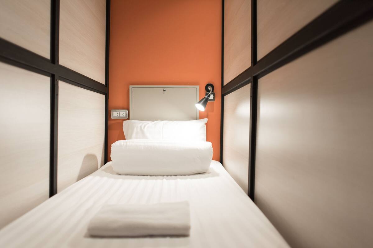 A single bed with white linens, a wall-mounted lamp, and a power outlet on the wall.