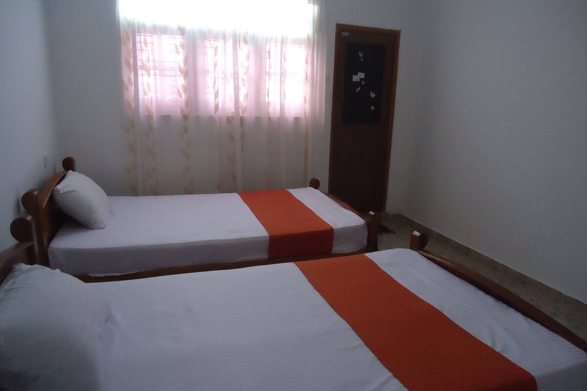 A room with two single beds, each with a wooden frame and white bedding. There is a wooden door with a small window, and a curtain with a leaf pattern.