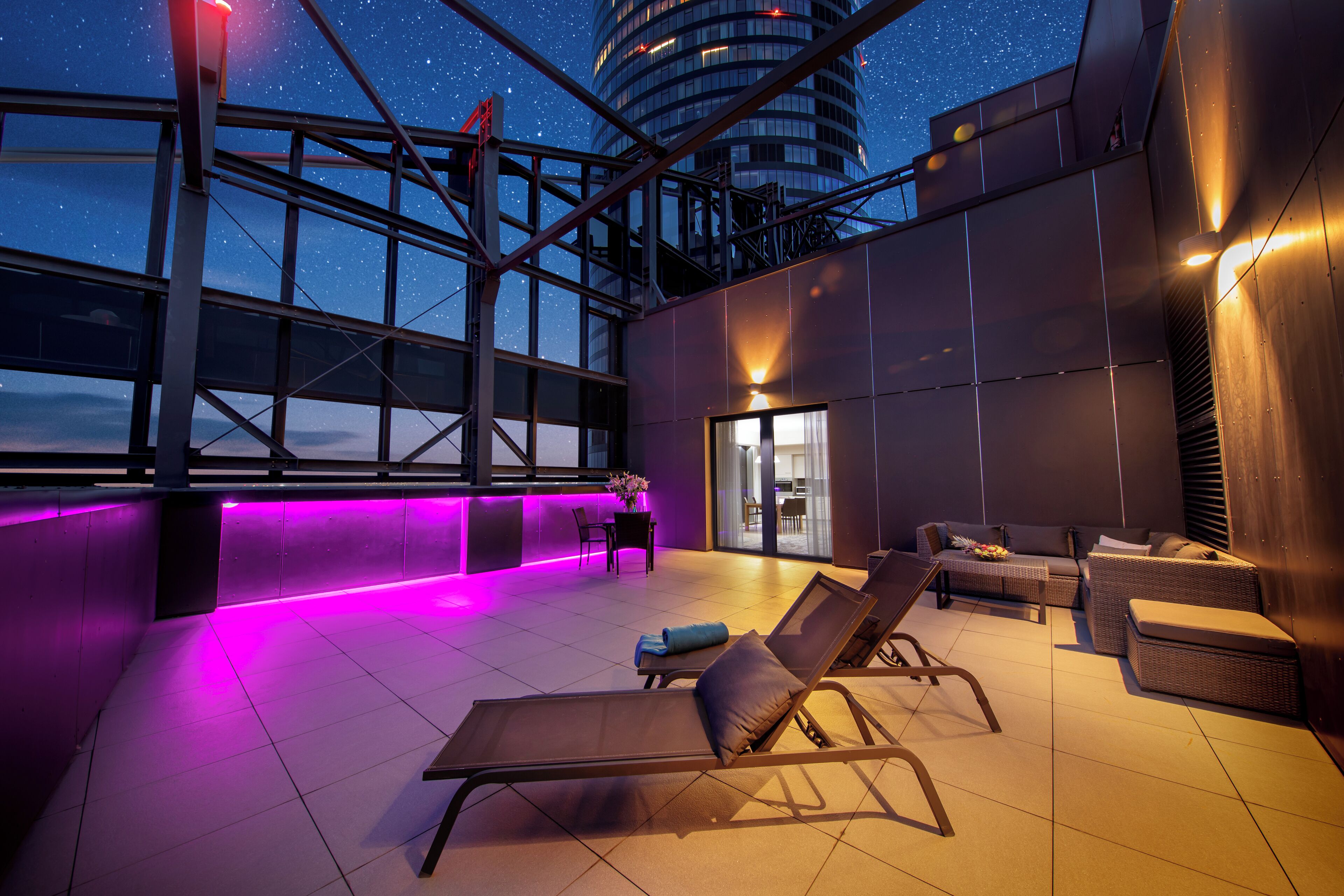A rooftop lounge with sun loungers, a table, and a view of the night sky.