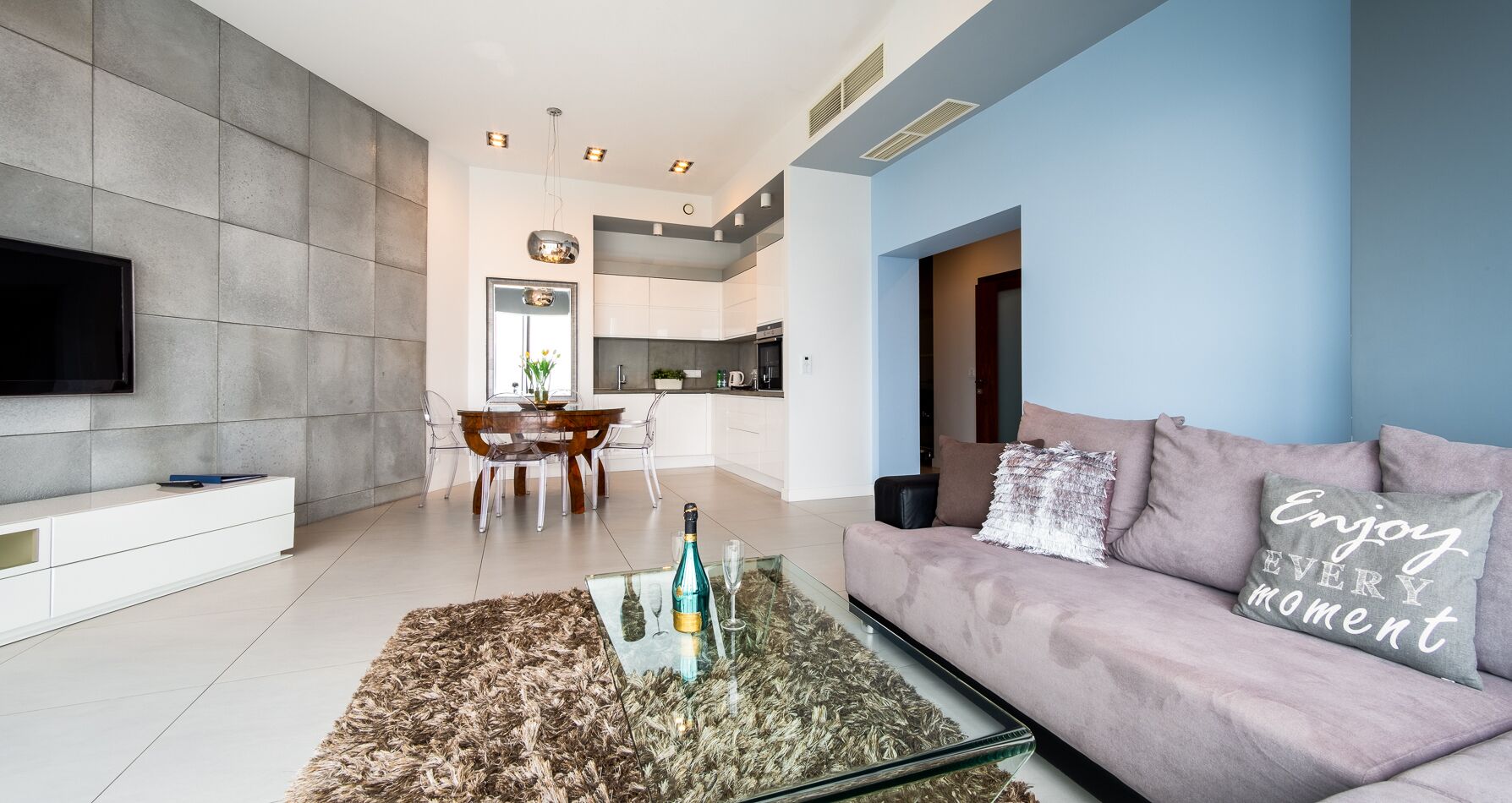 A modern living room with a glass coffee table, a sofa with a cushion, and a flat-screen TV mounted on the wall.