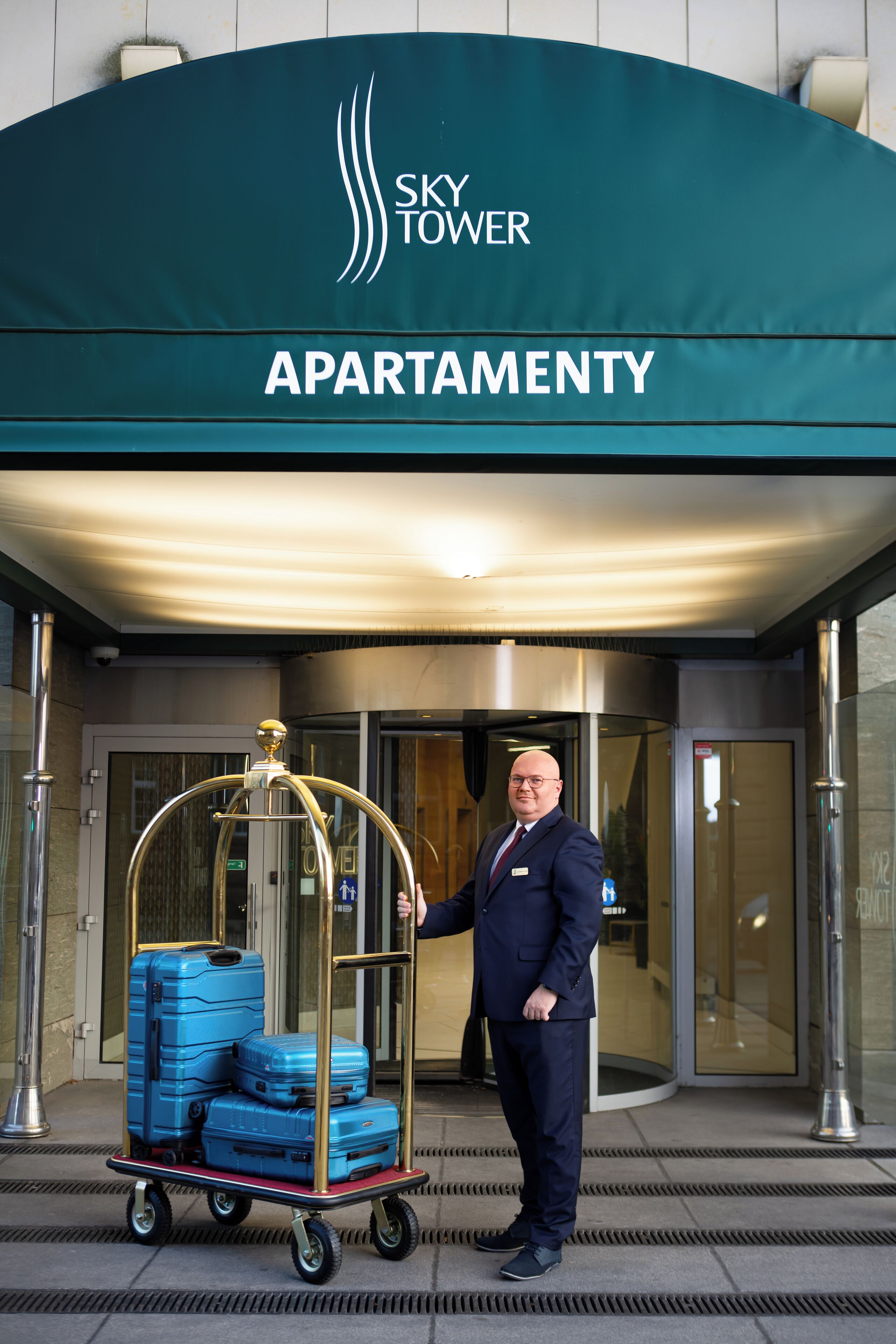 A man in a uniform standing in front of a hotel entrance with a luggage cart.