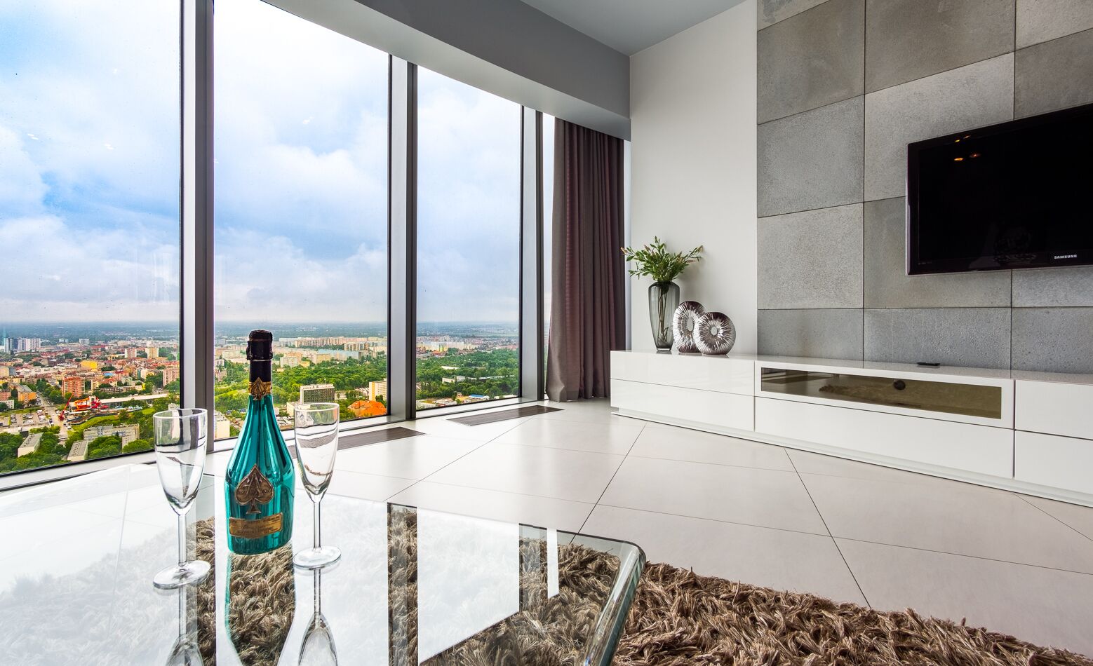 Ein modernes Wohnzimmer mit großem Fenster und Blick auf die Stadt, einem wandmontierten Flachbildfernseher sowie einem Glastisch mit Flasche und Gläsern.