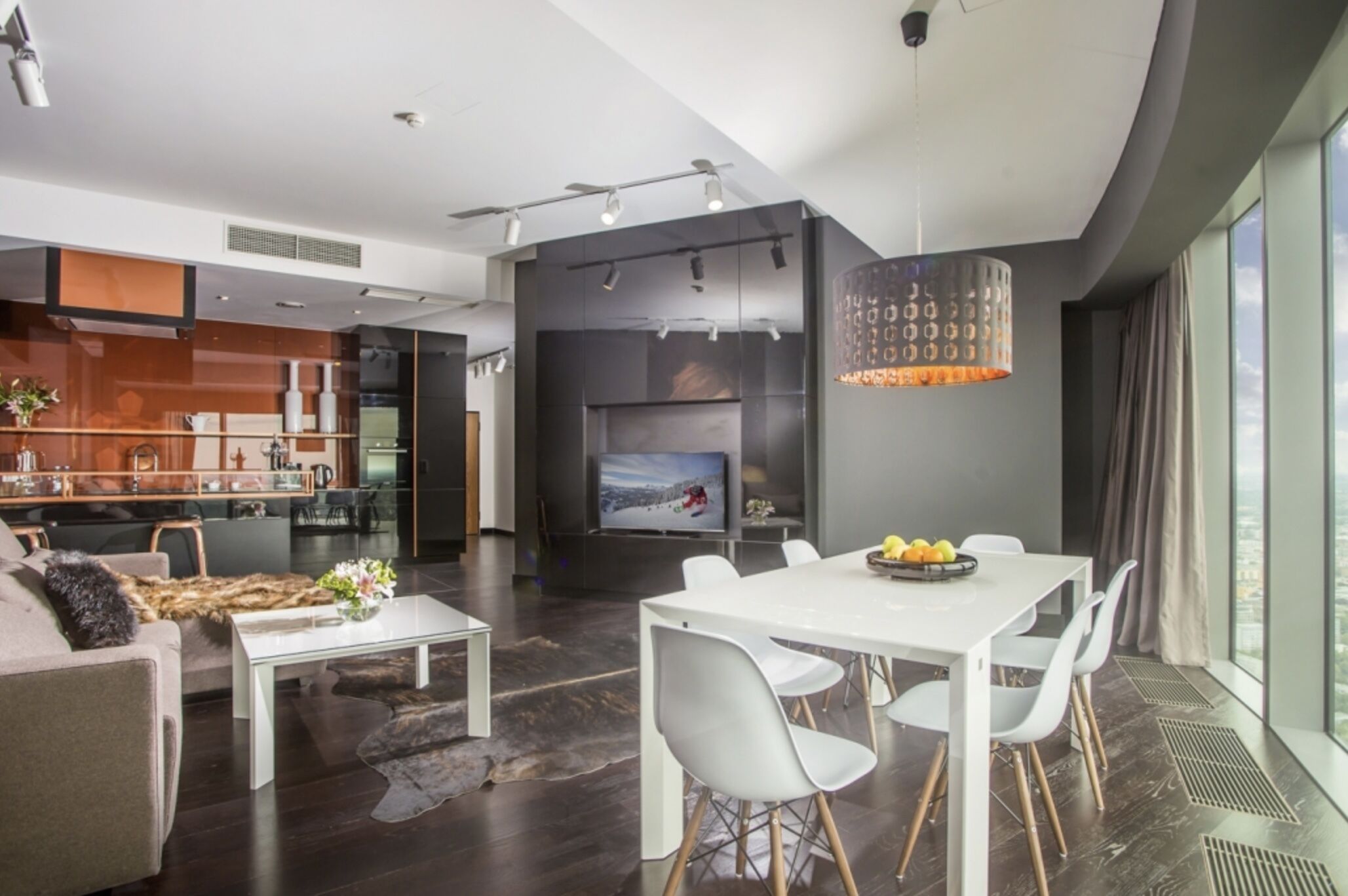 A modern living room with a dining area, a kitchen, and a large window providing a view of the city.