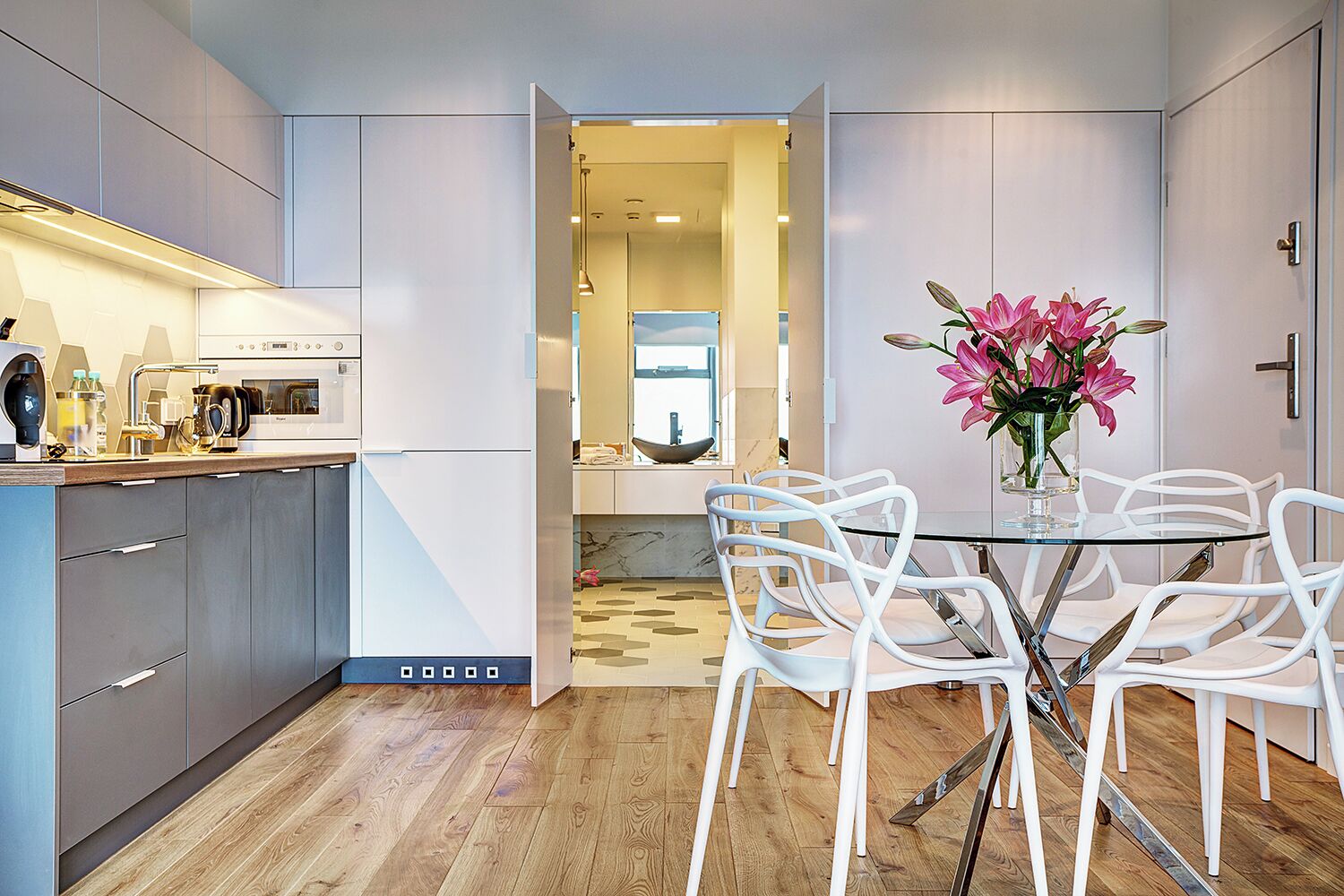 A modern kitchen with a built-in oven, a dining area with white chairs, and a vase of flowers on the table.