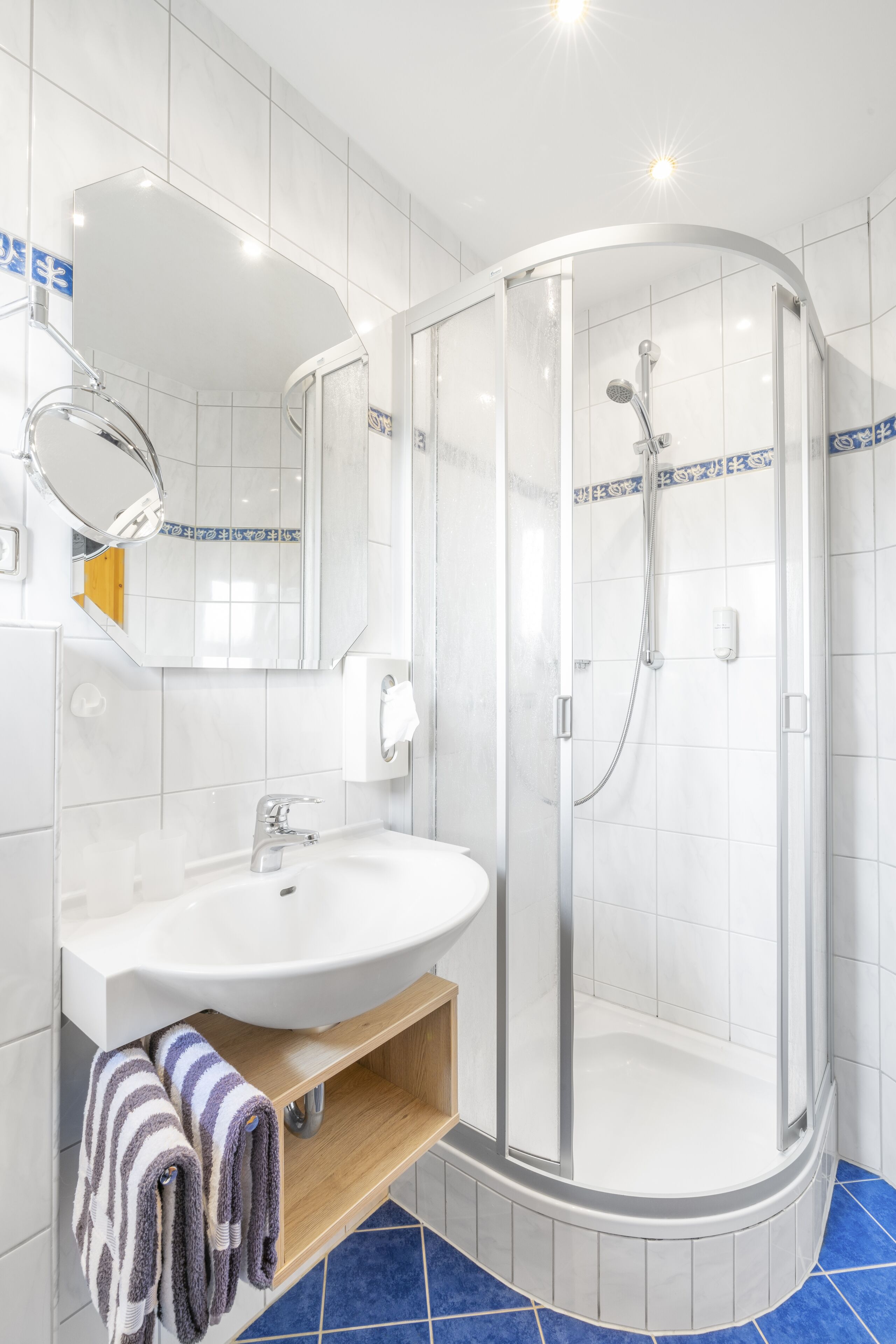 Un baño moderno con un lavamanos blanco, una ducha con mampara de vidrio y un suelo de baldosas azules.