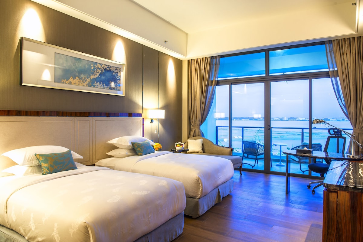 A hotel room with two beds, a desk, a chair, and a large window with a view of the sea.