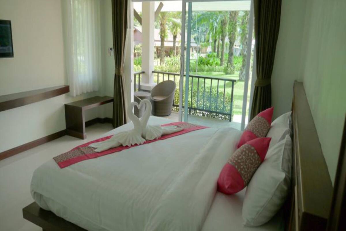 A hotel room with a bed, a bench, a chair, and a balcony with a view of greenery.