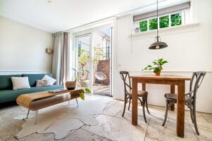 Uma sala de estar moderna com um sofá verde, um banco de madeira, uma mesa de jantar com cadeiras e uma janela grande com vista para uma parede de tijolos e vegetação lá fora.