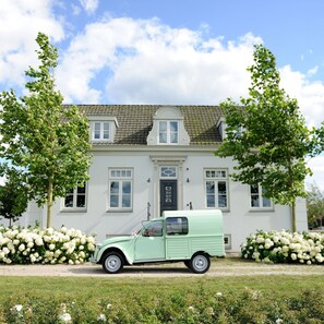 Une voiture verte d’époque est garée devant une villa blanche, dont le toit est recouvert de tuiles, et qui est entourée d’une végétation luxuriante et de fleurs blanches.
