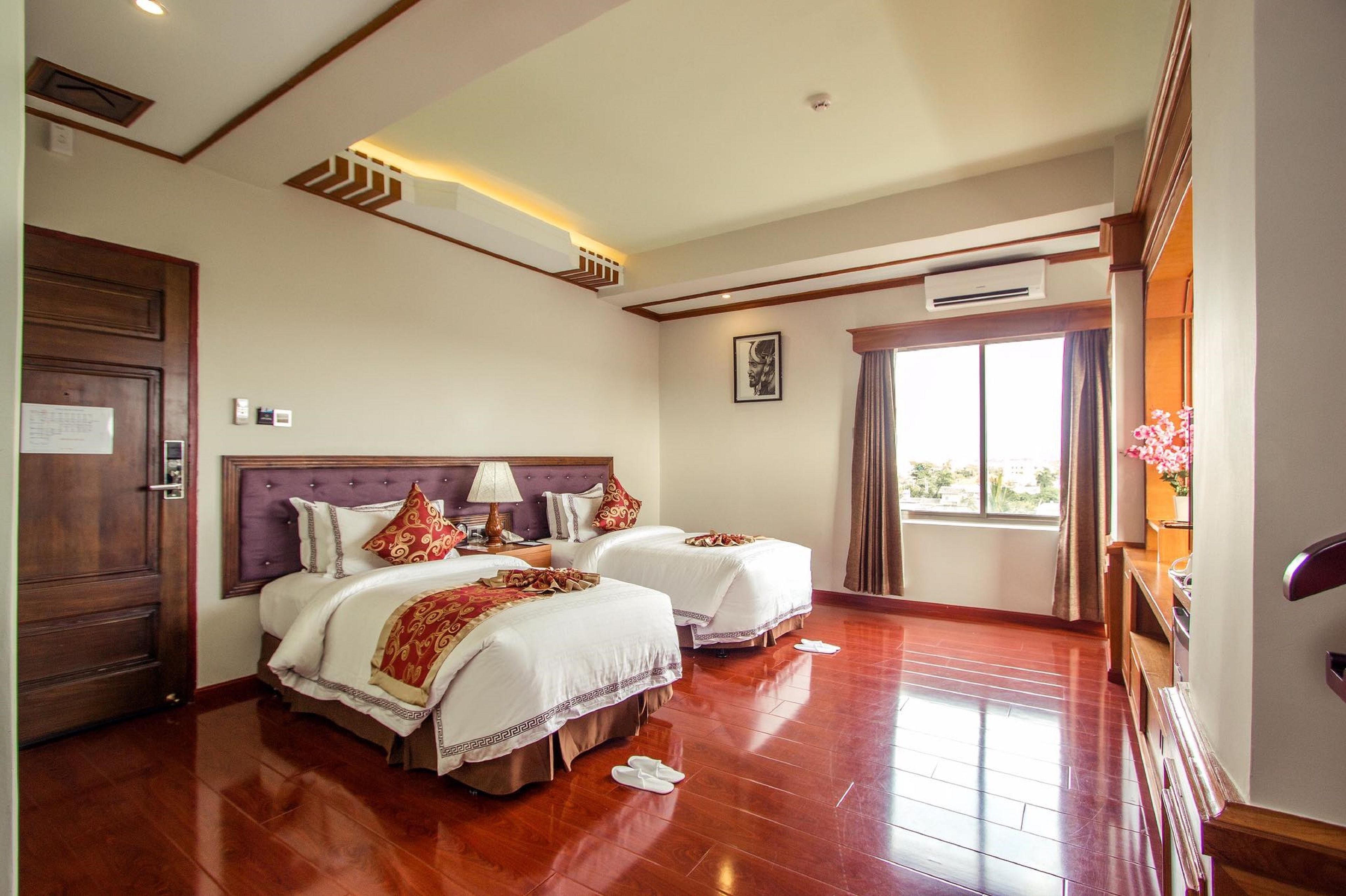 A hotel room with two beds, a wooden floor, a large window with curtains, and an air conditioning unit.