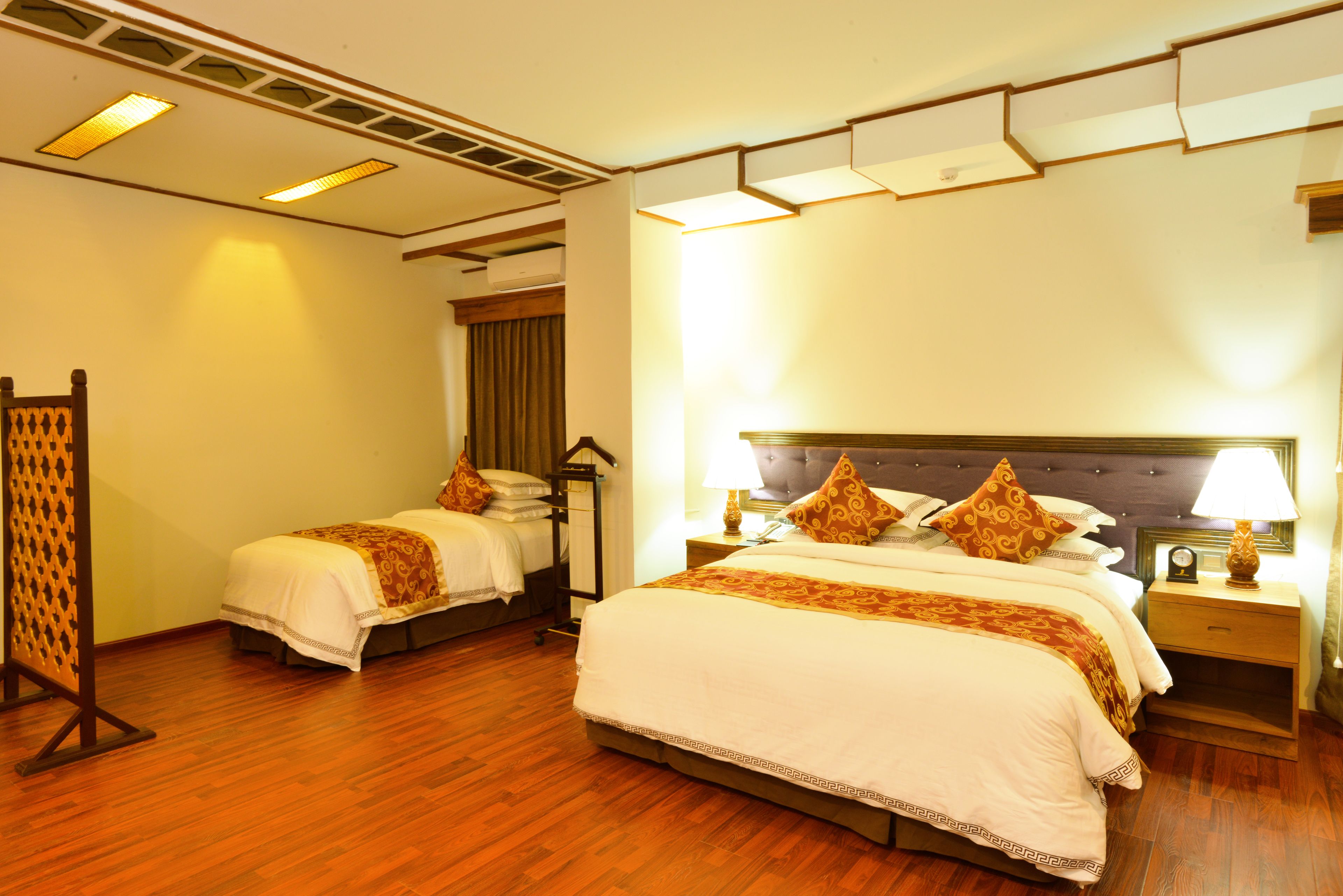 Habitación de hotel con dos camas individuales, suelo de madera, una cama grande con cabecera, mesitas de noche con lámparas y un panel decorativo en la pared.