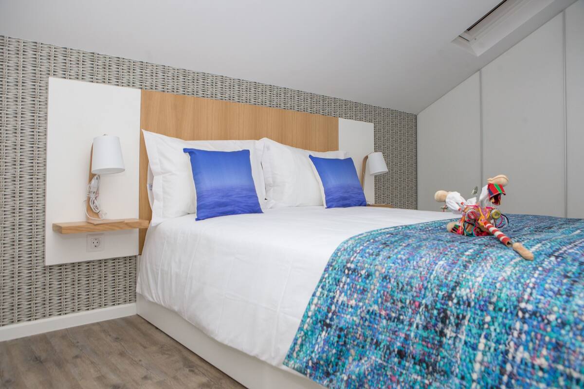 Une chambre d’hôtel moderne avec un grand lit, du linge de lit blanc, des oreillers bleus, une couverture colorée et un jouet sur le lit.