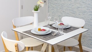 Una mesa de comedor dispuesta con sillas blancas, un jarrón con flores, y platos con comida y bebidas.