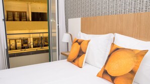 Habitación de hotel con cama, dos almohadas con diseños de frutas anaranjadas, una lámpara de noche y vista a un balcón con barandilla.
