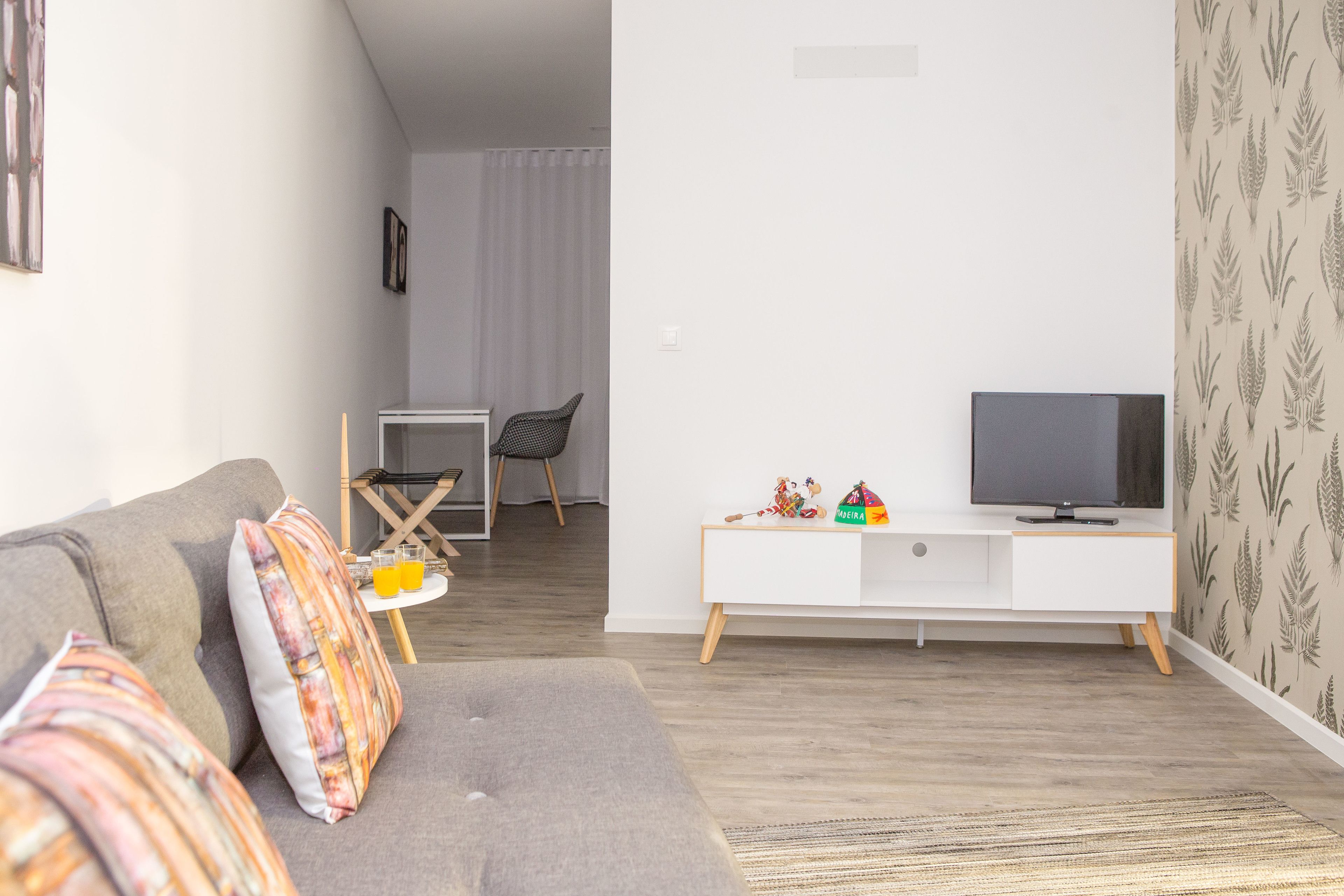 A living room with a grey sofa, a small white TV stand, and a patterned wallpaper.