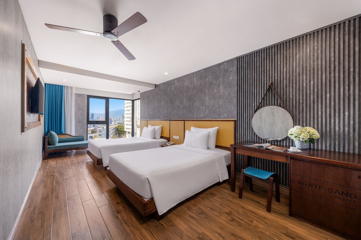 A hotel room with two beds, a desk, a chair, a ceiling fan, and a large window with a city view.