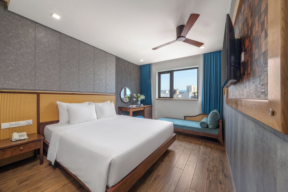 A hotel room with a large bed, a desk, a chair, a ceiling fan, a window with a view of buildings, and a mounted TV.