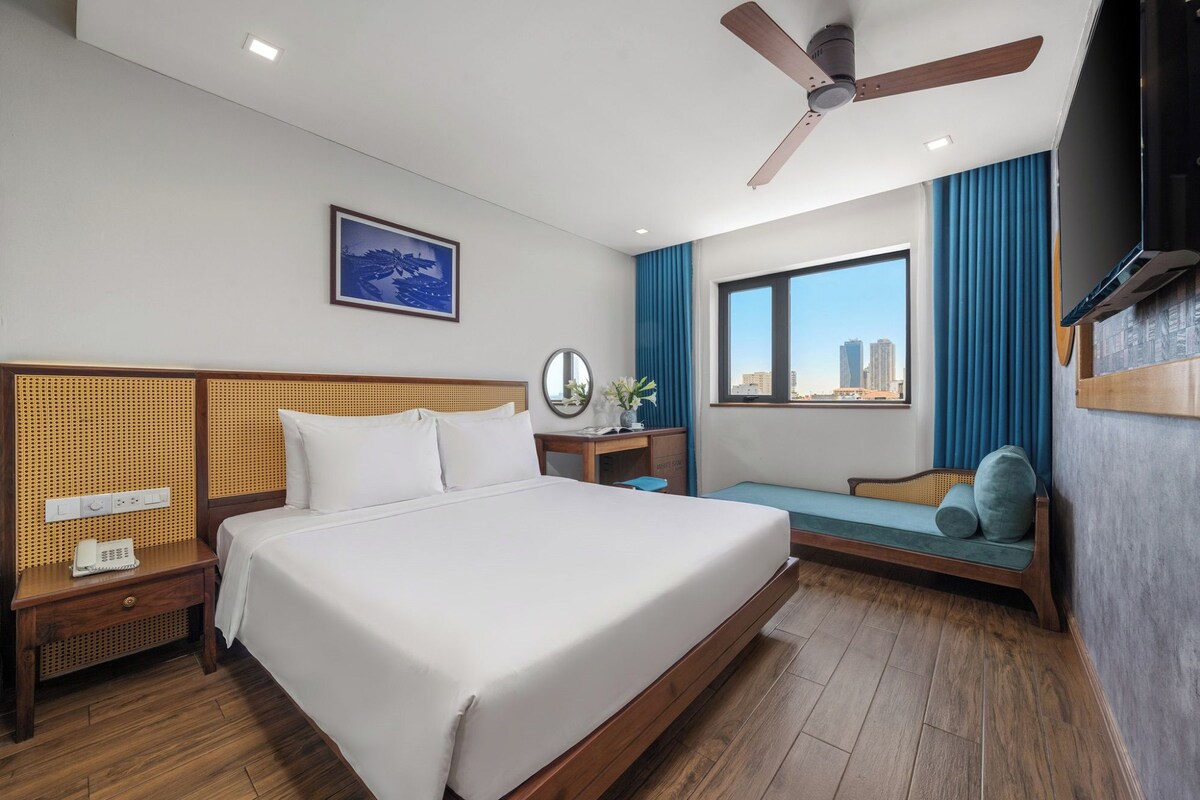 A hotel room with a large bed, a desk, a chair, a window with a city view, and a ceiling fan.