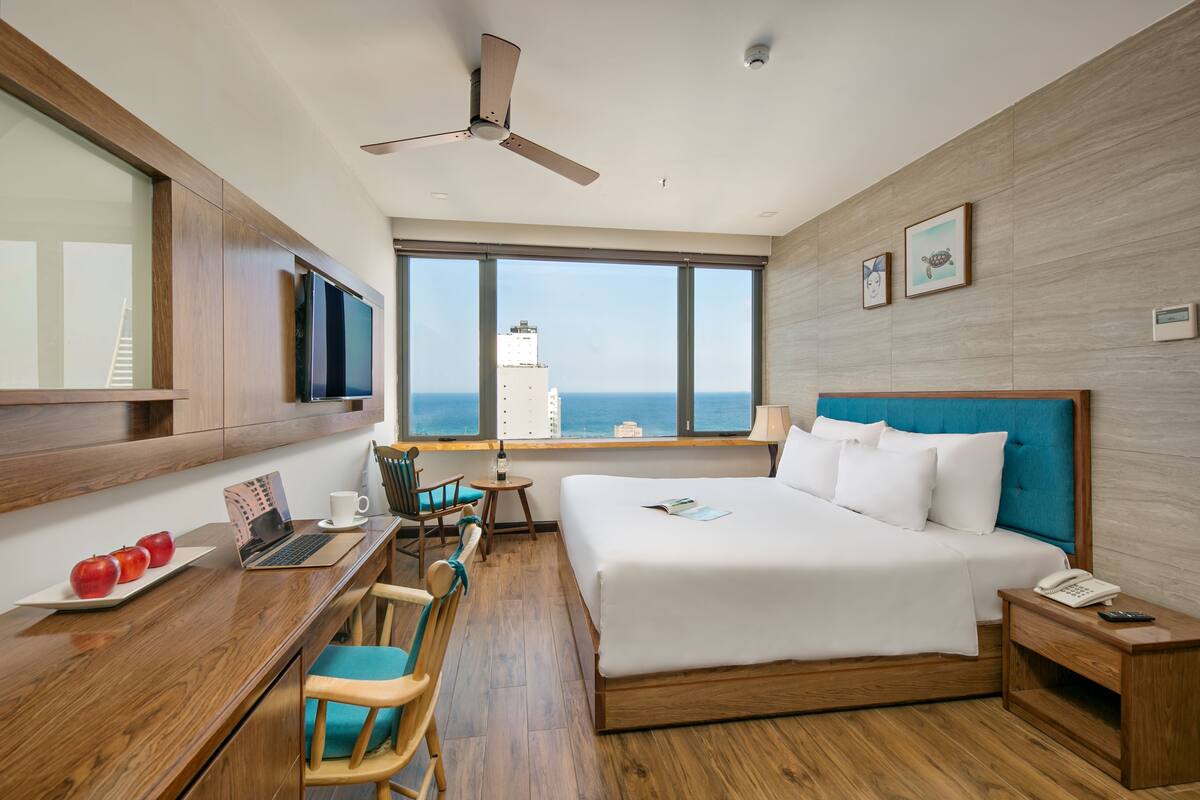Une chambre d’hôtel équipée d’un lit, d’un bureau, de chaises, d’une télévision, d’un ventilateur de plafond, d’une table de chevet et offrant une vue sur la mer.