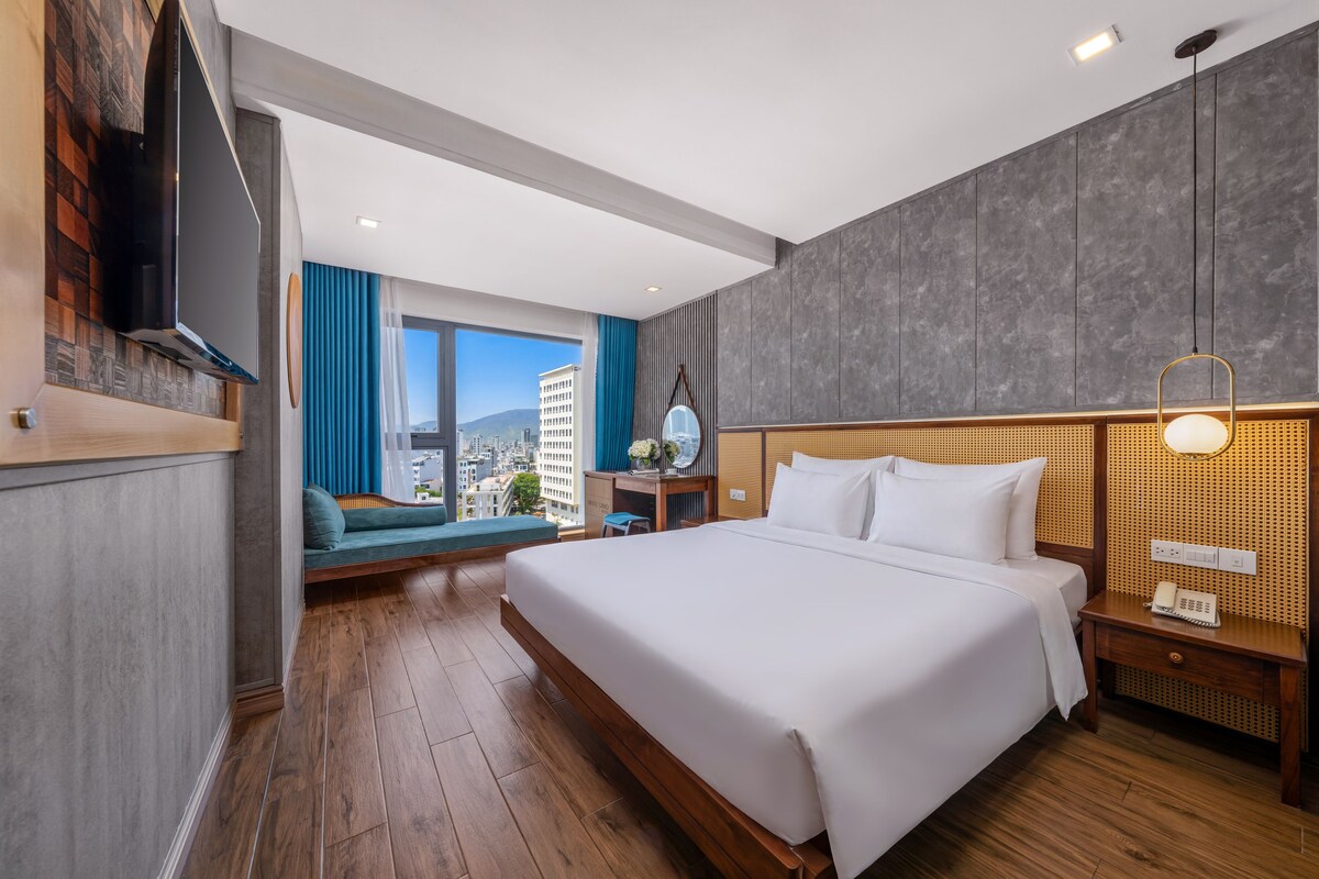 A hotel room with a large bed, a desk, a TV, and a view of the city.