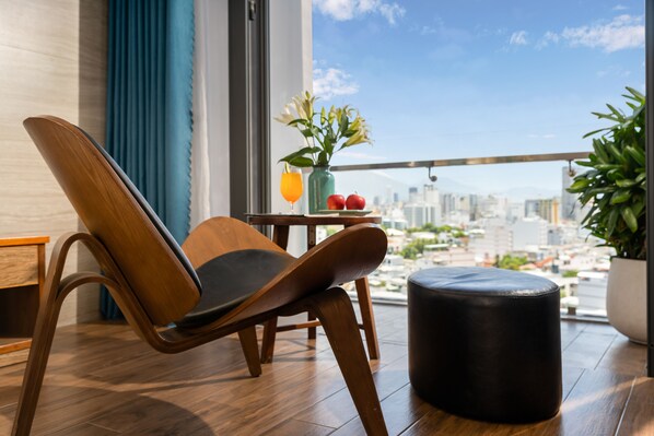 A modern room with a wooden chair, a small table with a vase of flowers, and a view of the city. - White Sand Boutique Hotel (Da Nang)