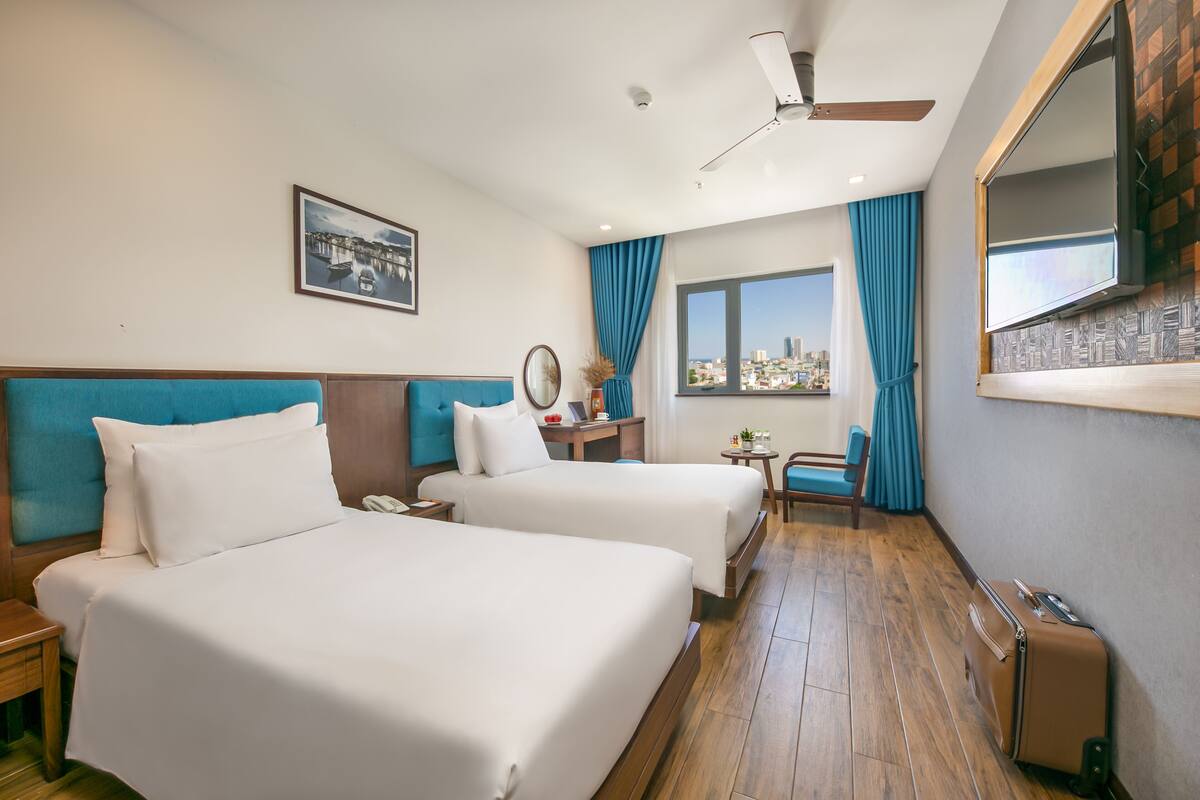 Une chambre d’hôtel avec deux lits, un bureau, une chaise, une fenêtre donnant sur la ville et un ventilateur de plafond.
