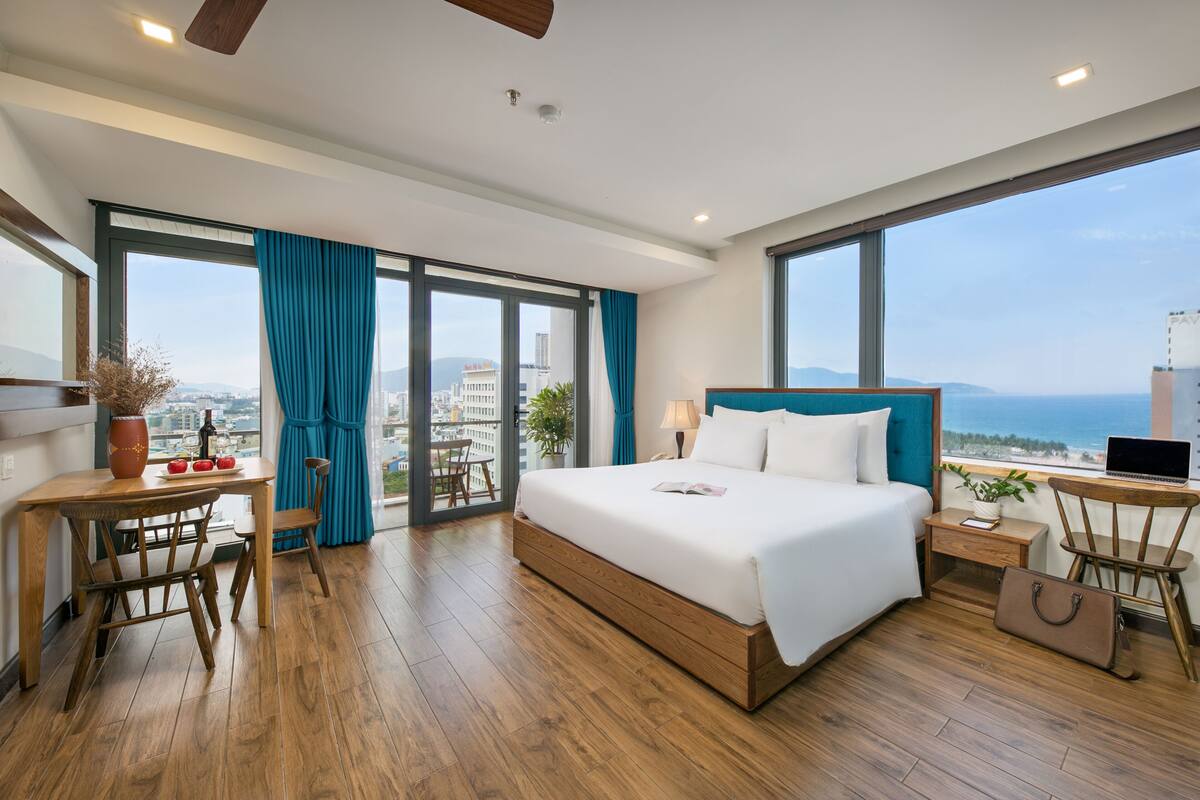 Une chambre d’hôtel moderne dotée d’un grand lit, d’un bureau avec un ordinateur portable, d’un coin repas et offrant une vue sur la ville et la mer.