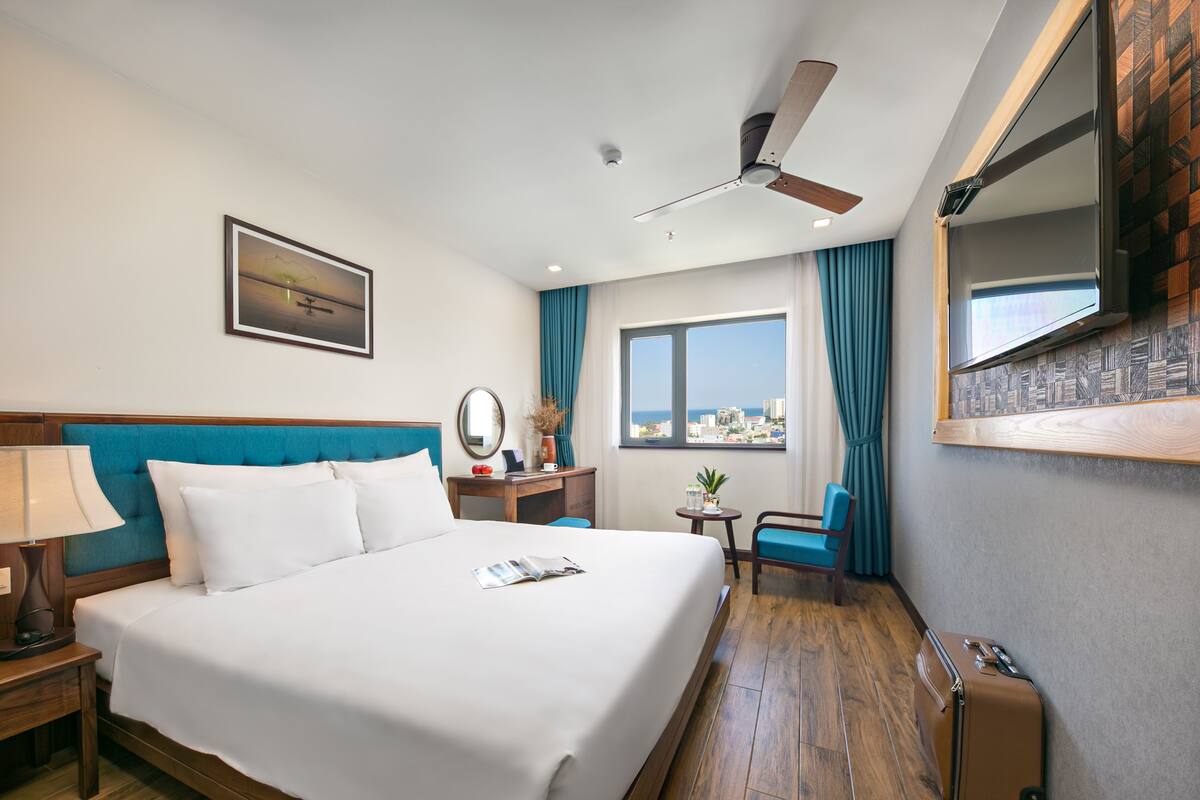 Une chambre d’hôtel avec un grand lit, un bureau, une chaise, un ventilateur de plafond, une fenêtre avec des rideaux et une vue sur la ville.