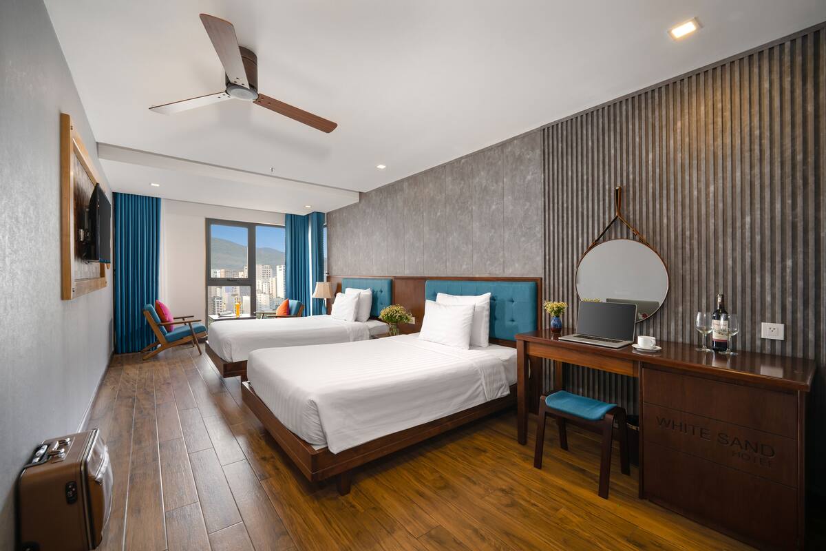 Une chambre d’hôtel avec deux lits, un bureau avec un ordinateur portable, une chaise, une valise et un ventilateur de plafond.