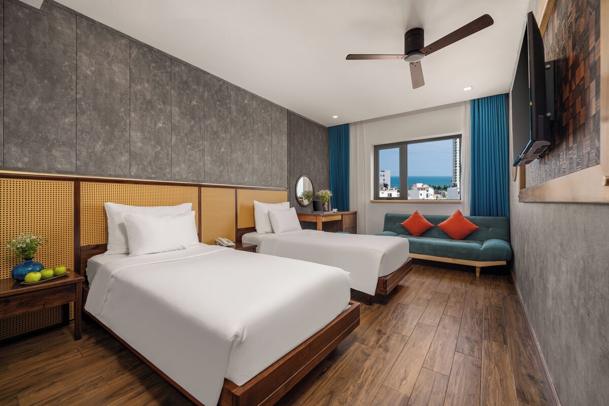 A hotel room with two beds, a sofa, a TV, and a view of the sea.