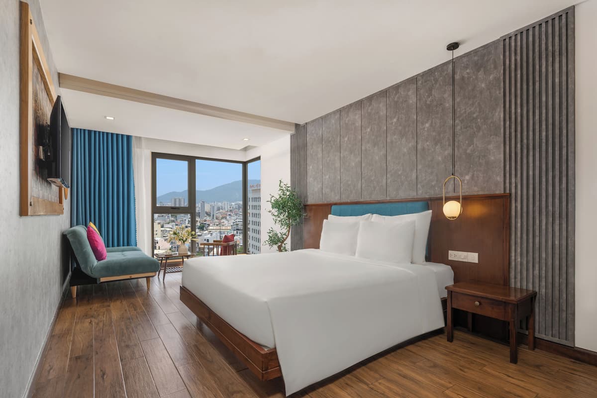 Une chambre d’hôtel moderne avec un grand lit, un coin salon comprenant un fauteuil et une petite table, et une vue sur la ville grâce à de grandes fenêtres.