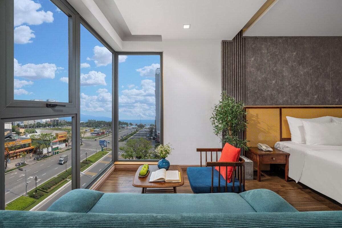 A modern hotel room with a large window, a bed, a desk, and a view of the city.