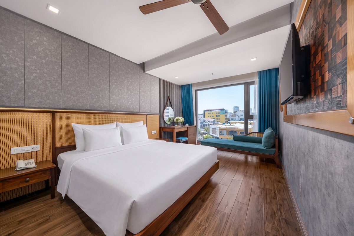 A hotel room with a large bed, wooden headboard, a desk with a chair, a flat-screen TV mounted on the wall, and a view of the city through the window.