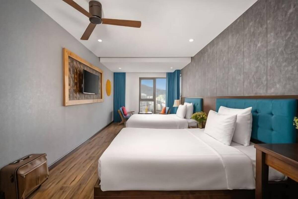 Une chambre d’hôtel avec deux lits, un ventilateur de plafond, une télévision, une fenêtre avec des rideaux et une vue sur la ville.