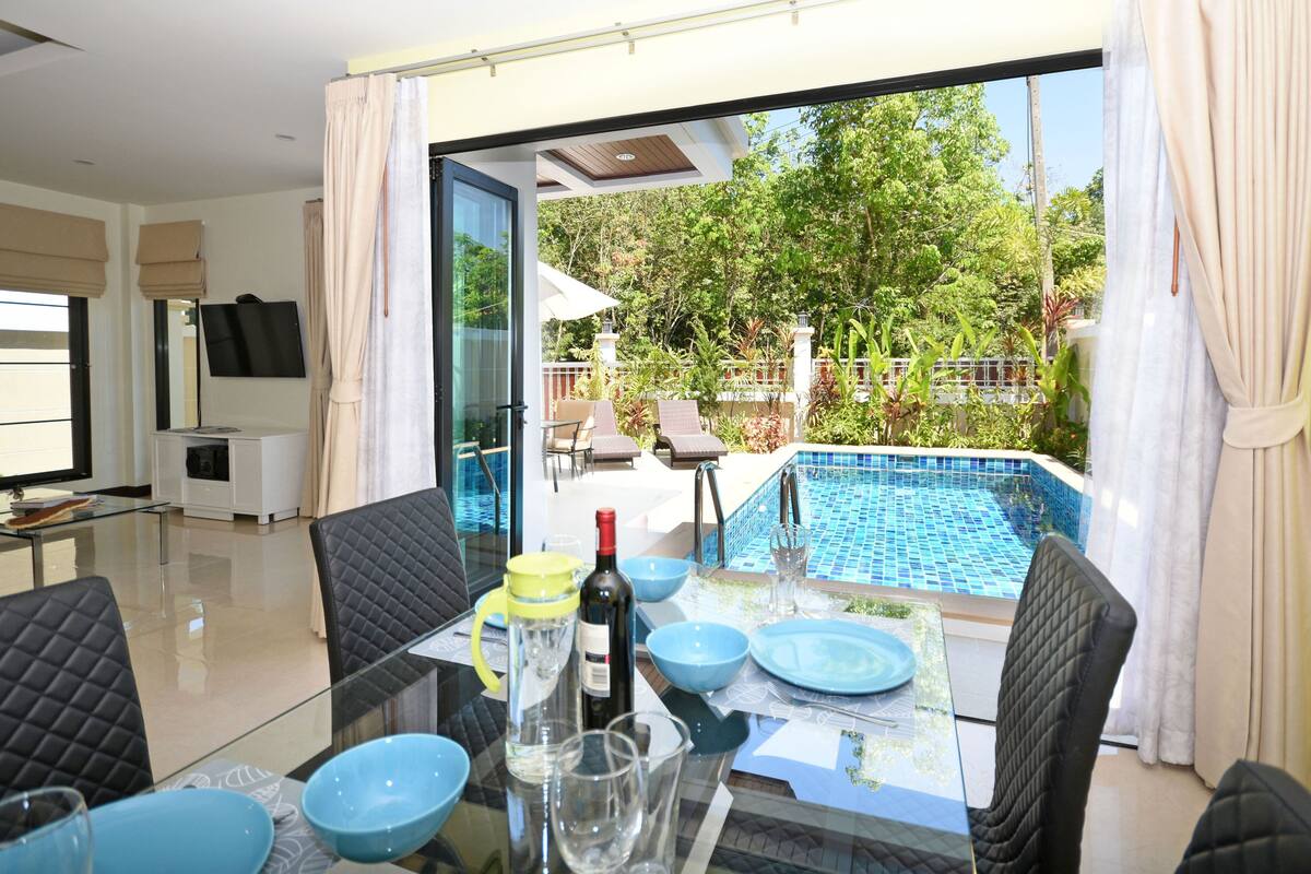 A dining area with a glass table, blue bowls, and chairs. A TV is mounted on the wall, and a pool is visible through the glass doors.