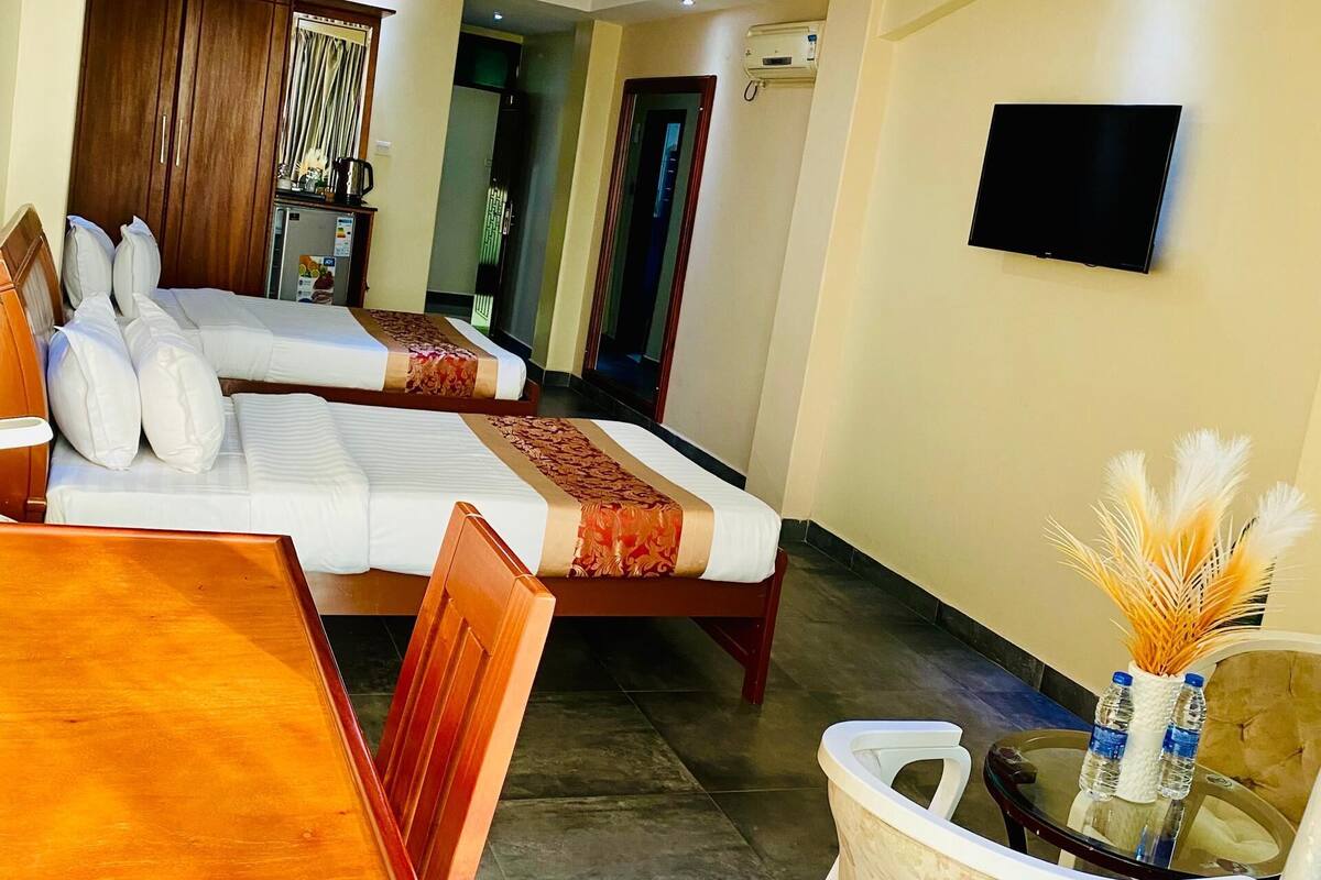 A hotel room with two beds, a dining table, a TV, and a small kitchenette.