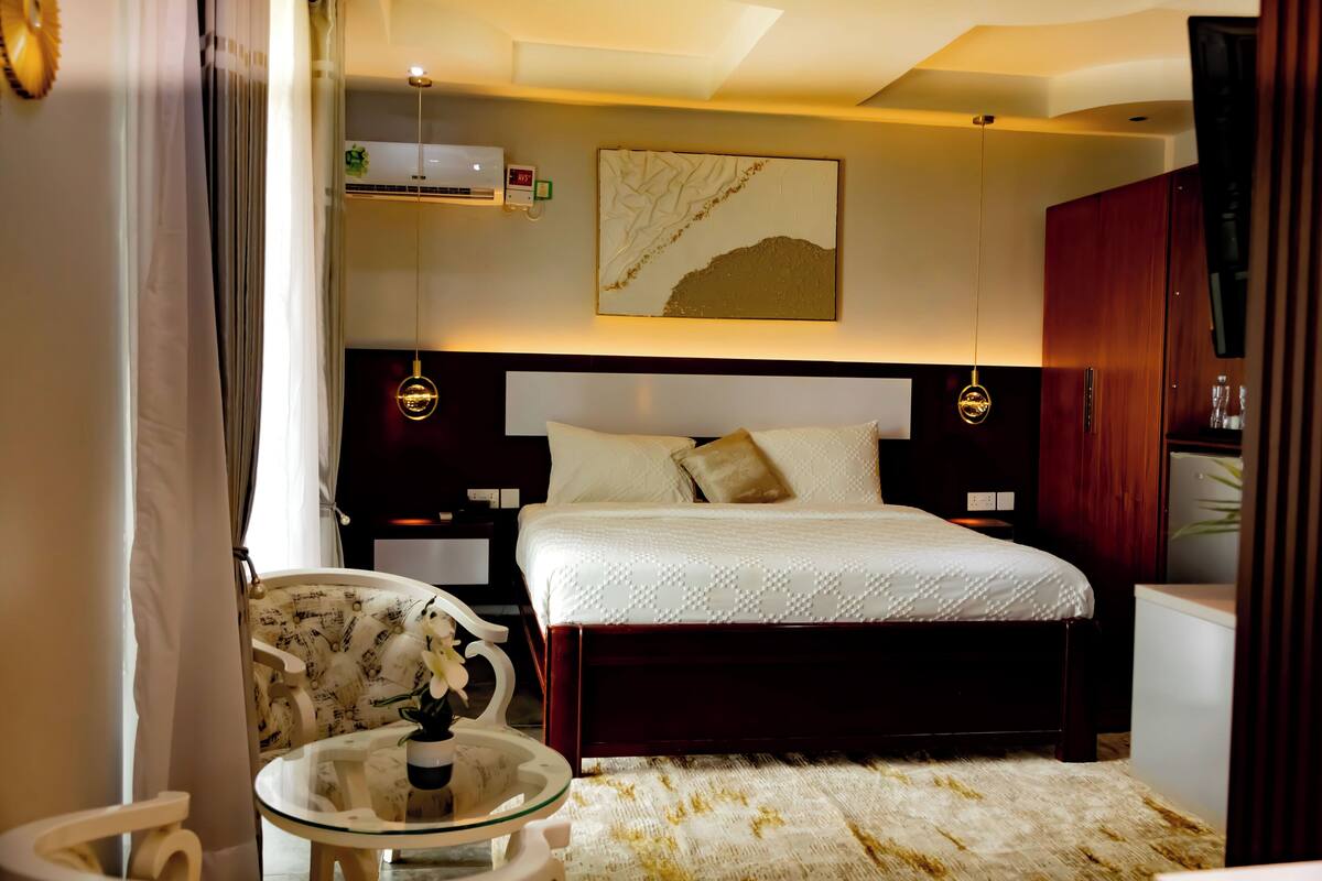 A hotel room with a bed, bedside tables, a chair, a small table with a vase, a large abstract painting, and a ceiling light fixture.