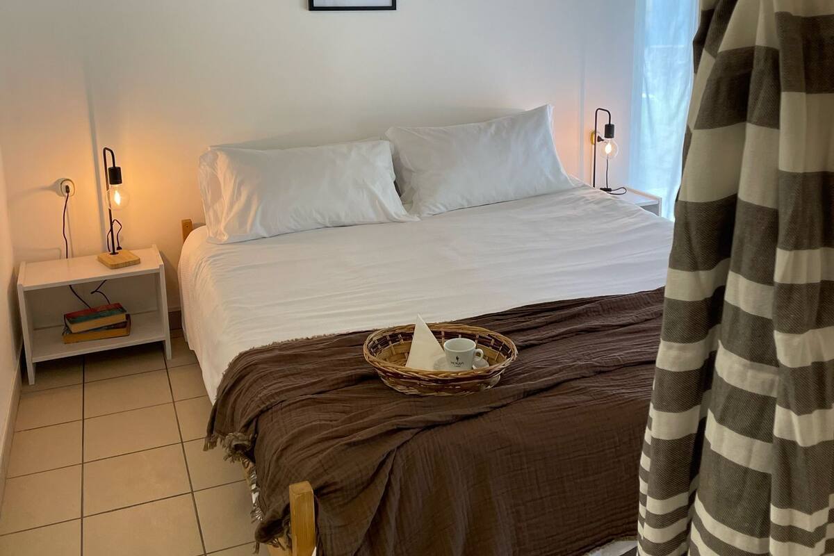 A bedroom with a bed, two bedside tables, a wall-mounted light fixture, a framed picture, and a window with a view.