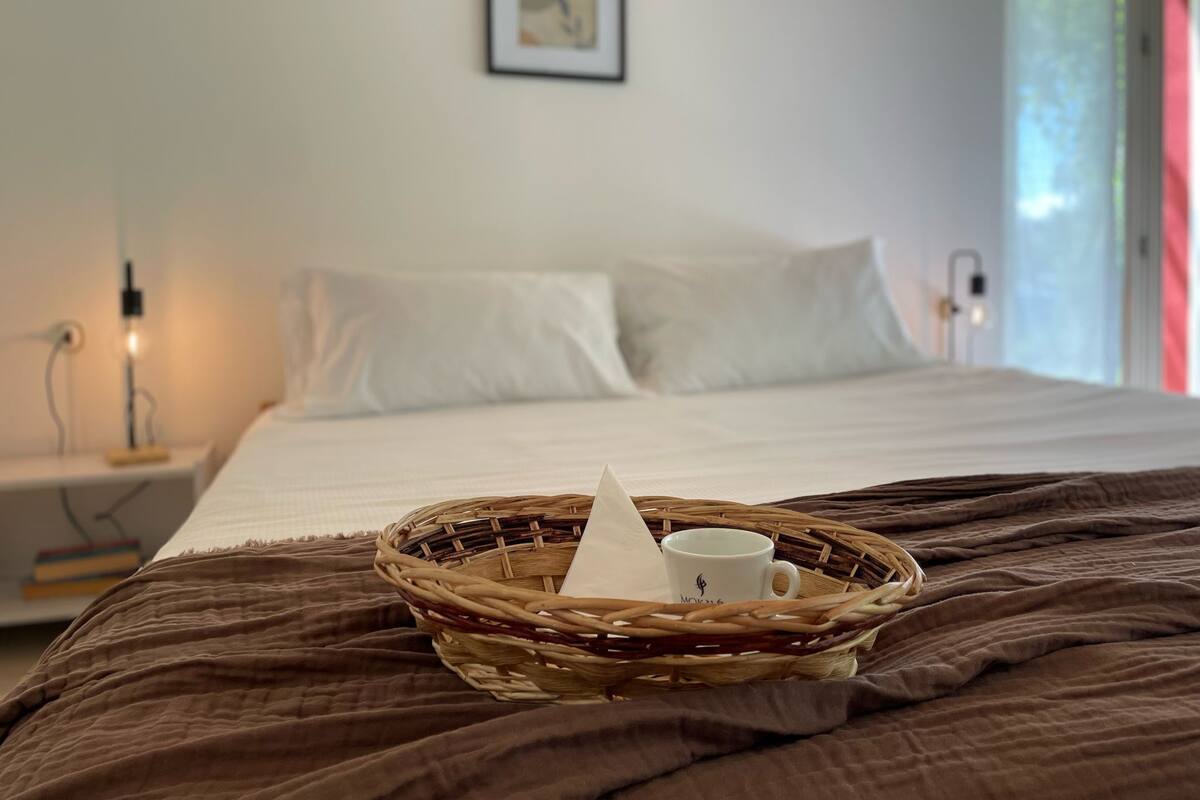 A neatly made bed with a brown blanket, a white pillow, and a white pillow on the headboard. A wicker basket with a white mug and a white napkin is placed on the bed.