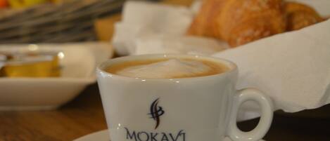 A cup of coffee labeled "MOKAVI CAFE" on a saucer, with croissants and fruits in the background.